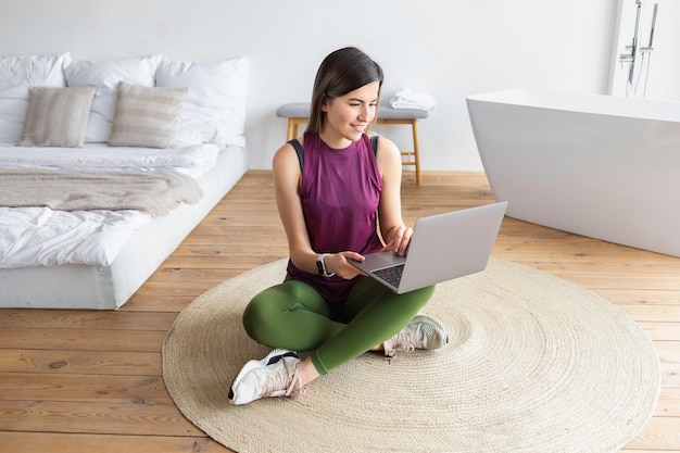 Deportiva mujer caucásica joven sentada en el suelo en casa utiliza la computadora portátil para ver el entrenamiento en línea. Concepto de estilo de vida saludable