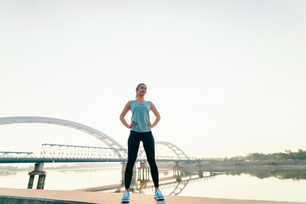 Deportiva mujer caucásica de pie en el kay con las manos en las caderas en la mañana. En el puente de fondo. Longitud total.