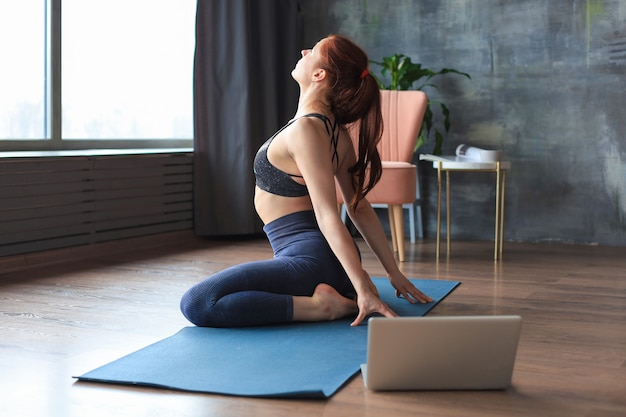 Deportiva mujer delgada practicando yoga avanzado frente a la computadora portátil, viendo tutoriales en línea. Estilo de vida saludable.