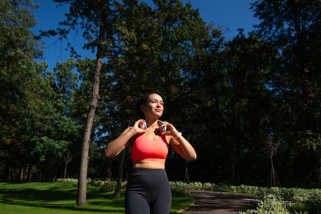 Deportiva mujer joven con auriculares en el cuello caminando sobre una carrera en el parque después de hacer ejercicio al aire libre