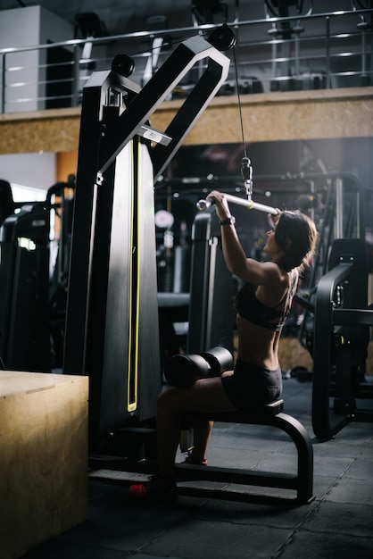 Deportiva mujer joven con cuerpo atlético perfecto vistiendo ropa deportiva negra con simulador de entrenamiento para bombear los músculos de la espalda en el gimnasio. Concepto de estilo de vida saludable y entrenamientos en un moderno gimnasio oscuro.