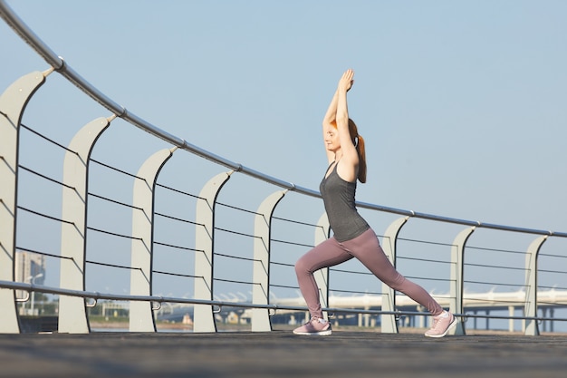 Deportiva mujer de mediana edad con los ojos cerrados realizando pose de guerrero con los brazos levantados en el terraplén de la ciudad