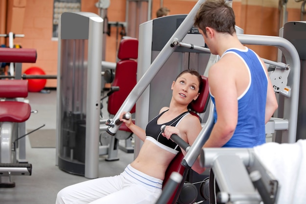 Deportiva mujer sentada en una prensa de hombro hablando con su entrenador en un gimnasio