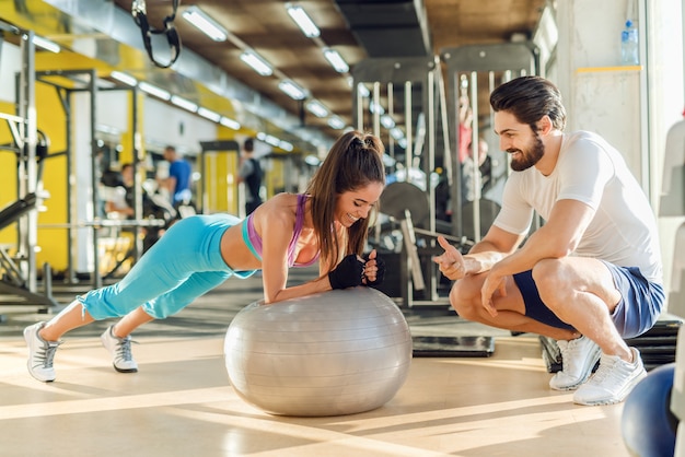 Deportiva mujer sonriente haciendo tablas de pilates mientras su entrenador personal se agacha junto a ella y la anima.