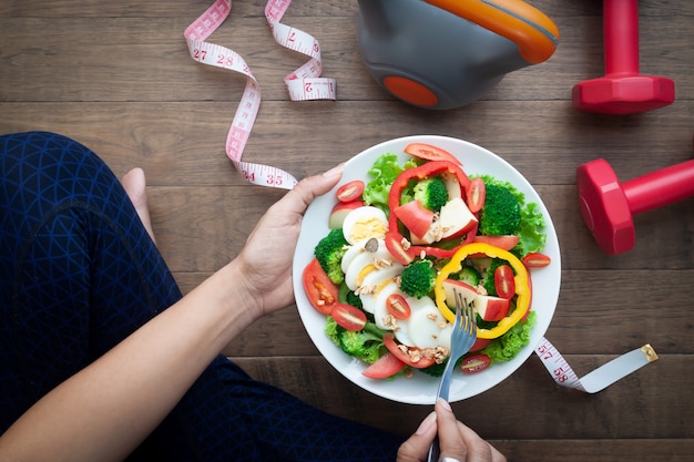 Deportiva mujer sosteniendo plato de ensalada con equipos de deporte en el piso de madera