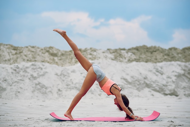 Deportiva mujer de yoga en la playa practica patadas hacia atrás apoyándose en sus manos. Concepto de deporte y estilo de vida saludable.