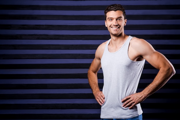 Deportivo y guapo. Joven deportivo en la parte superior del tanque tomados de la mano en la cadera y sonriendo mientras está de pie contra el fondo de rayas