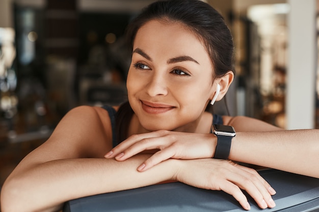 Deportivo y hermoso. Retrato de mujer linda y alegre con reloj inteligente en su mano mirando a otro lado mientras descansa en el gimnasio. Concepto de deporte y fitness. Estilo de vida saludable. Digital