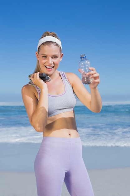 Foto deportivo rubia feliz de pie en la playa con una botella de agua y saltar la cuerda