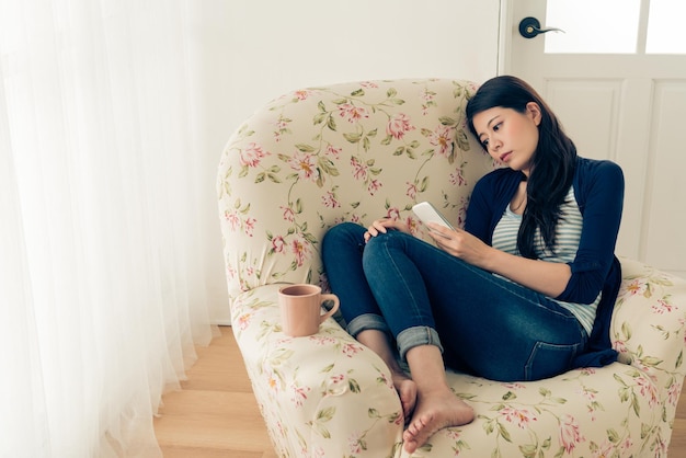 Depression Junges Mädchen, das Probleme hat, auf dem Sofa vor dem Wohnzimmerfenster zu sitzen und auf das Mobiltelefon zu schauen und auf eine Nachricht zu warten. Vintage Retro-Filmfarbe.