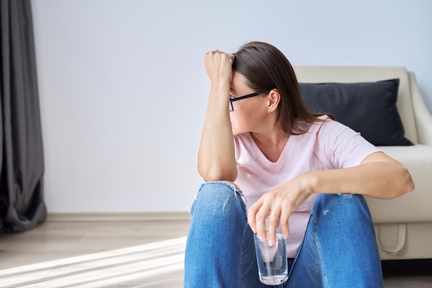 Depressionen, Neurosen, Ängste, Stress, psychische Probleme, die Folgen der Krankheit Coronavirus Covid-19. Traurige, unglückliche, reife Frau, die zu Hause mit einem Glas Wasser auf dem Boden sitzt