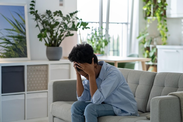 Depressive, erschöpfte, unglückliche junge Frau sitzt auf der heimischen Couch und versteckt das Gesicht in den Händen, fühlt sich allein betrogen