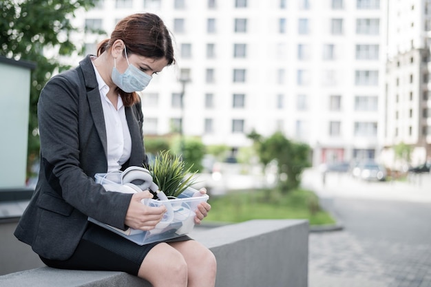 Depressive Frau mit medizinischer Maske sitzt auf Brüstung im Freien mit einer Kiste persönlicher Gegenstände vom Schreibtisch. Arbeitsloses Mädchen allein auf der Straße. Insolvenz eines Unternehmens aufgrund von Quarantäne im Coronavirus