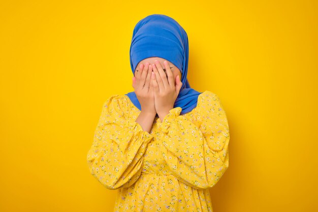 Depressive junge Asiatin in Freizeitkleidung, die das Gesicht mit der Hand bedeckt und große Probleme isoliert auf gelbem Hintergrund hat