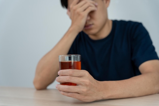Depressiver junger asiatischer Mann, der süchtig ist und sich schlecht fühlt, allein zu Hause Whisky trinkt, gestresst, frustriert, einsam, Alkohol trinkt, unter problematischem Alkoholismus, Alkoholismus, Lebens- und Familienproblemen leidet