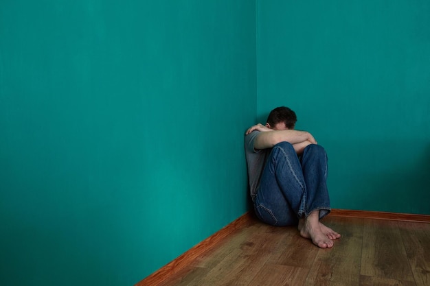 Depressiver Mann sitzt in einer Ecke in einem leeren grünen Raum mit dem Kopf im Schoß. Platz kopieren.