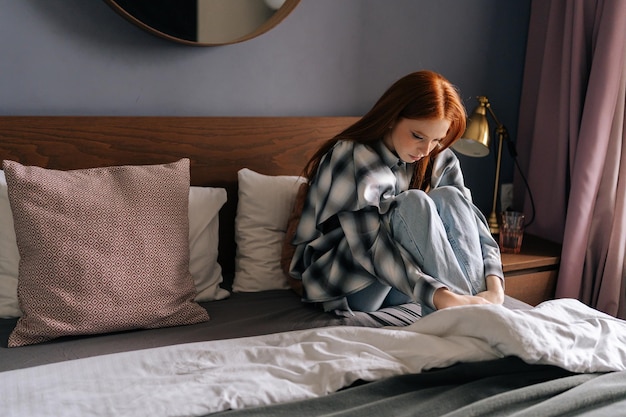 Deprimierte junge Frau sitzt allein auf dem Bett und umarmt Knie im Schlafzimmer vor dem Hintergrund des Sonnenlichts aus dem Fenster. Verzweifelte hübsche Frau fühlt Enttäuschung