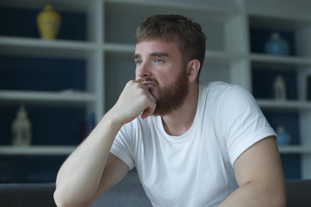 Deprimierter, unglücklicher, trauriger Mann mittleren Alters, der auf der Couch im Wohnzimmer sitzt und sich auf seine Hände stützt