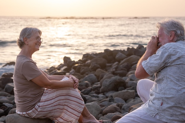 Der ältere Mann fotografiert seine Frau, die bei Sonnenuntergang am Strand sitzt