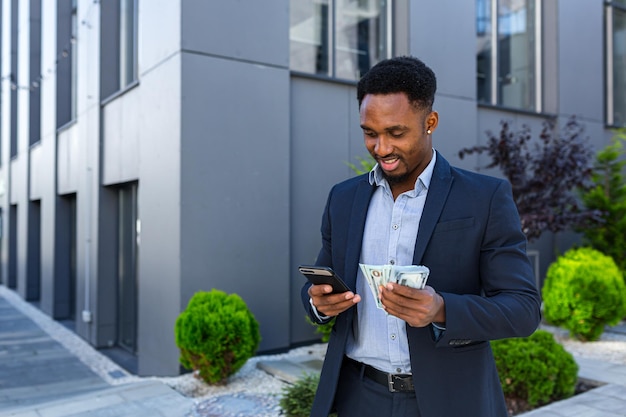 Der afroamerikanische Geschäftsmann, der auf dem modernen Gebäude der Straße steht, zeigt die Gewinne beim Wetten von Bargeld mit dem Handy in den Händen. schwarzer Mann im Anzug im Freien mit Smartphone und Packdollar