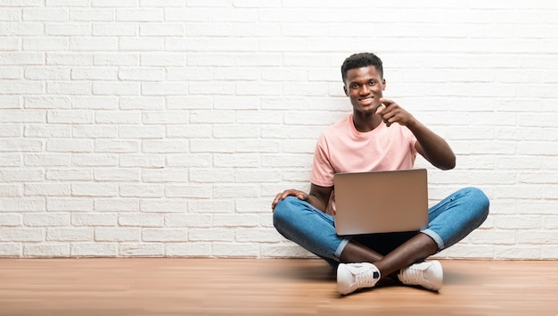 Der afroamerikanische Mann, der auf dem Boden mit seinem Laptop sitzt, zeigt Finger auf Sie