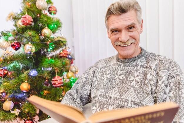 Der alte Mann hält ein Buch neben dem Weihnachtsbaum