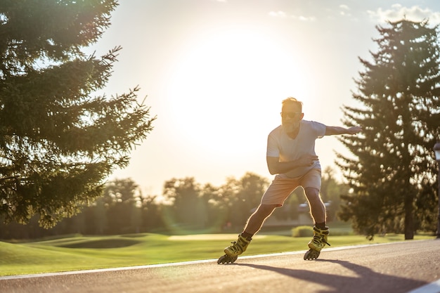 Der alte Mann mit Sonnenbrille beim Rollerblading auf dem Sonnenuntergangshintergrund