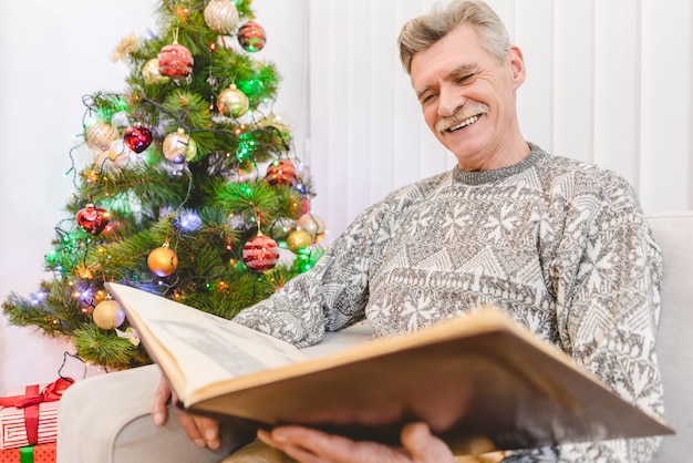 Der alte Mann schaut in ein Fotoalbum neben dem Weihnachtsbaum