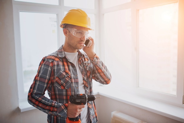 Der Arbeiter oder Baumeister hält eine Bohrmaschine und telefoniert. Bau- oder Reparaturkonzept. Vor dem Hintergrund des Bauens