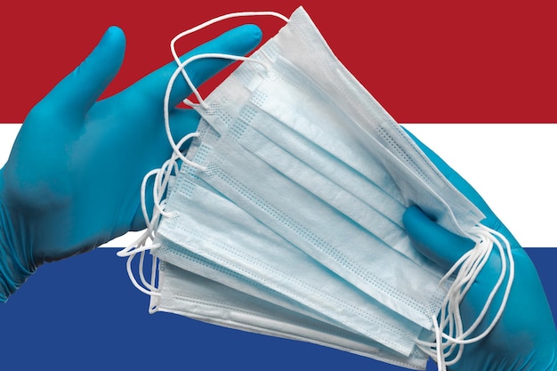 Der Arzt hält Gesichtsmasken in den Händen blaue Handschuhe auf der Hintergrund-Nationalflagge der Niederlande. Konzept Quarantäne, Hygiene, Pandemieausbruch. Chirurgischer antibakterieller Gesichtsverband