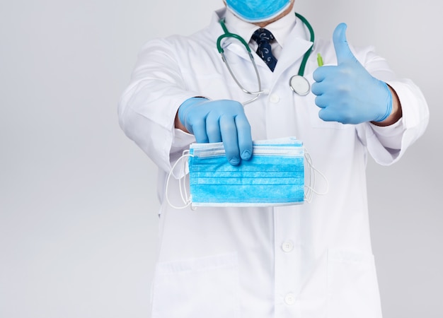Der Arzt in einem weißen Kittel und sterilen blauen Latexhandschuhen hält medizinische Textilmasken in der Hand