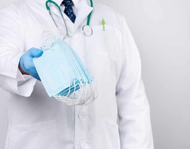 Der Arzt in einem weißen Kittel und sterilen blauen Latexhandschuhen hält medizinische Textilmasken in der Hand