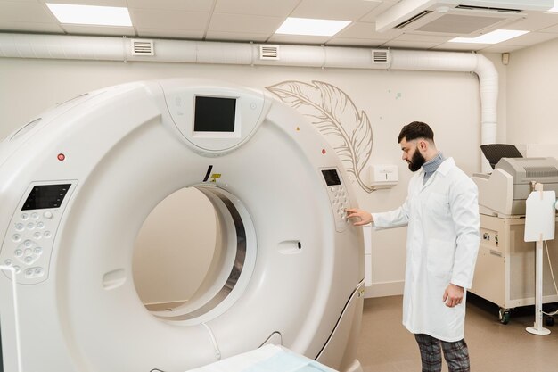 Der Arzt stellt den CT-Computertomographen in der medizinischen Klinik ein Gutaussehender bärtiger Radiologe in medizinischer Robe mit Computertomographen, um detaillierte innere Bilder des Körpers des Patienten zu erhalten