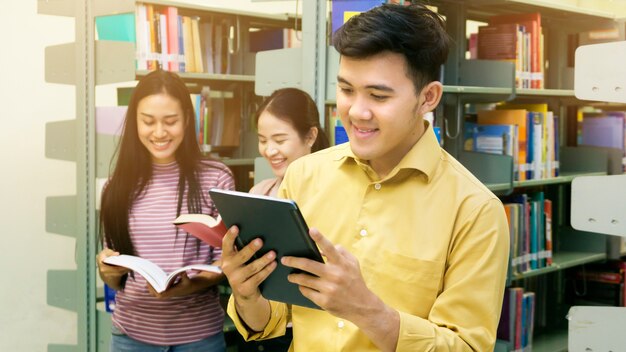 der asiatische Jugendlichmann liest eine Tablette mit Mädchen sprechen am Bücherregal