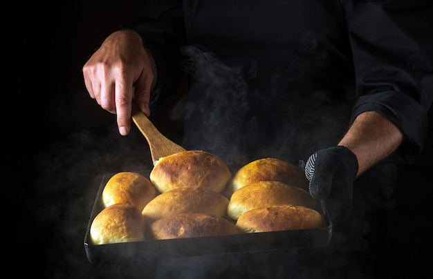 Der Bäcker dreht die Kuchen auf einem heißen Backblech um Das Konzept der Herstellung von Hamburgerbrötchen Schwarzer Platz für Menü oder Rezept