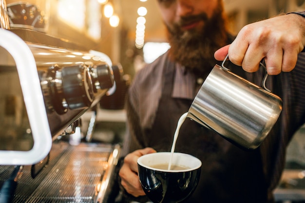 Der bärtige Barista gießt Milch mit Kaffee in die Tasse.