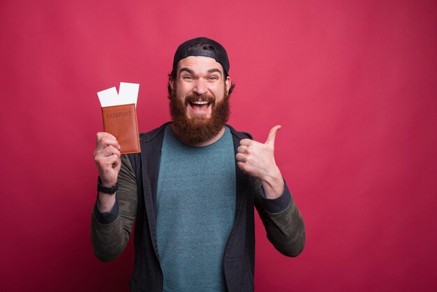 Der bärtige Hipster zeigt den Daumen nach oben, während er seinen Pass mit Tickets hält