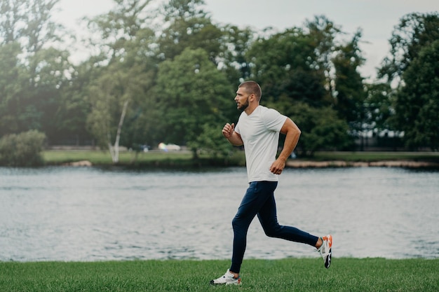 Der bärtige Mann des Athleten läuft entlang einer wunderschönen Landschaft, demonstriert Ausdauer in aktiver Kleidung, geht in der Nähe des Flusses joggen und atmet frische Luft Sport, der Fitness- und Trainingskonzept ausübt
