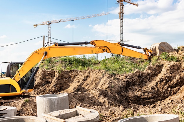 Der Bagger gräbt eine Baugrube für den Einbau von Abwasserbrunnen. Turmdrehkrane im Hintergrund. Aushubarbeiten - Versorgung von Wohngebäuden mit Wasser und Abwasser.