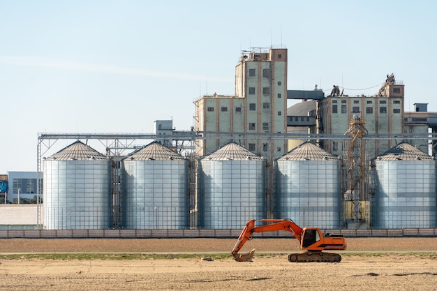 Der Bagger steht vor dem Hintergrund Silbersilos Agro-Produktionsanlage für die Verarbeitung Trocknung Reinigung und Lagerung von landwirtschaftlichen Produkten Große Eisenfässer Getreidespeicher Aufzug