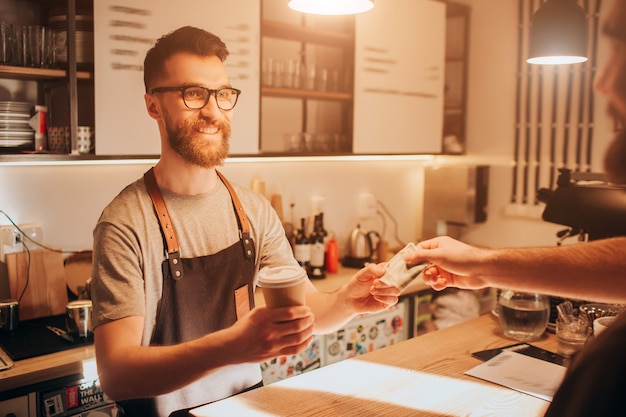 Der barkeeper steht hinter der bar und hält eine tasse kaffee, die er für den kunden gemacht hat. der barmann sieht glücklich und lächelnd aus, während er vom kunden eine bestellung in bar erhält.