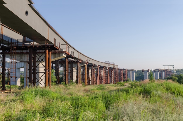 Der Bau einer Brücke über den Fluss mit den Stützen, Konstruktionselementen, Kränen