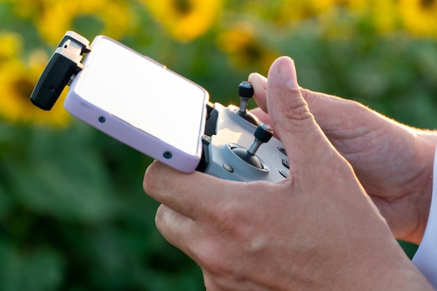 Der Bediener hält die Fernbedienung der Drohne vor dem Hintergrund eines Sonnenblumenfeldes und Wolken in den Händen Ein Bauer in einem weißen T-Shirt und einer Kappe verwendet einen Quadcopter, um in einer Industrie zu arbeiten