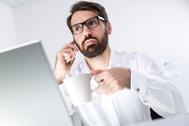 Der beschäftigte junge Mann in einem weißen Hemd trägt eine Brille und hält seine Kaffeetasse, während er auf seinem Telefon spricht und in die Ferne schaut.