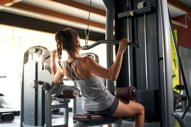 Der Blick von hinten auf eine junge Frau in Sportshorts und ein graues Tanktop, das im Fitnessstudio fit ist, gibt einem speziellen Simulator die Hand