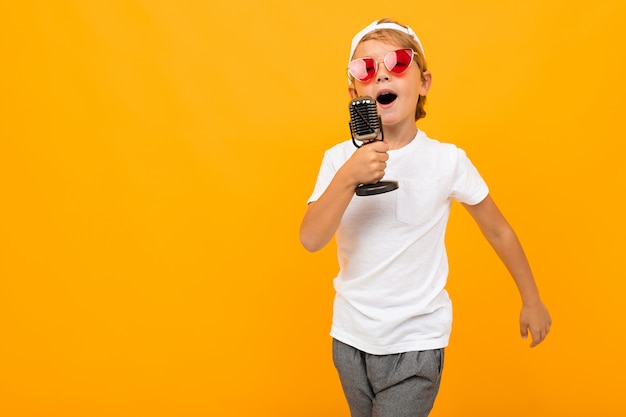 Der blonde Junge singt in ein Mikrofon an einer orangefarbenen Wand