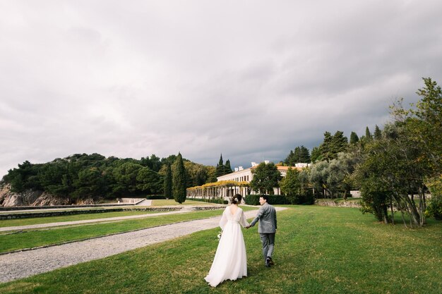 Foto der bräutigam blickt zurück auf die braut, die ihm entlang des grünen rasens der villa milocer montenegro folgt.