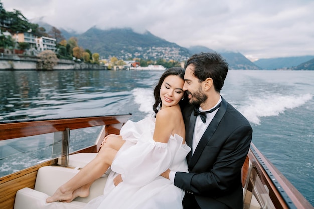 Der Bräutigam umarmt die Braut von hinten, lehnt sich gegen ihn, sitzt auf einer Yacht, die auf dem Lake Como in Italien segelt