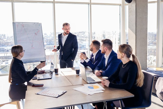 Der Chef steht mit Grafiken neben der Tafel, führt Statistiken vor, verschiedene Mitarbeiter nehmen an der Schulung teil, stellt die neuen Produkte des Unternehmens vor, berichtet über die Arbeitsergebnisse für Partner.