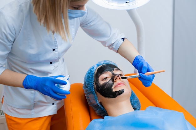 Der Dermatologe schmiert eine schwarze Maske auf das Gesicht, um die Laser zu verjüngen und das Kohlenstoff-Peeling durchzuführen. Dermatologie und Kosmetologie. Mit einem chirurgischen Laser.